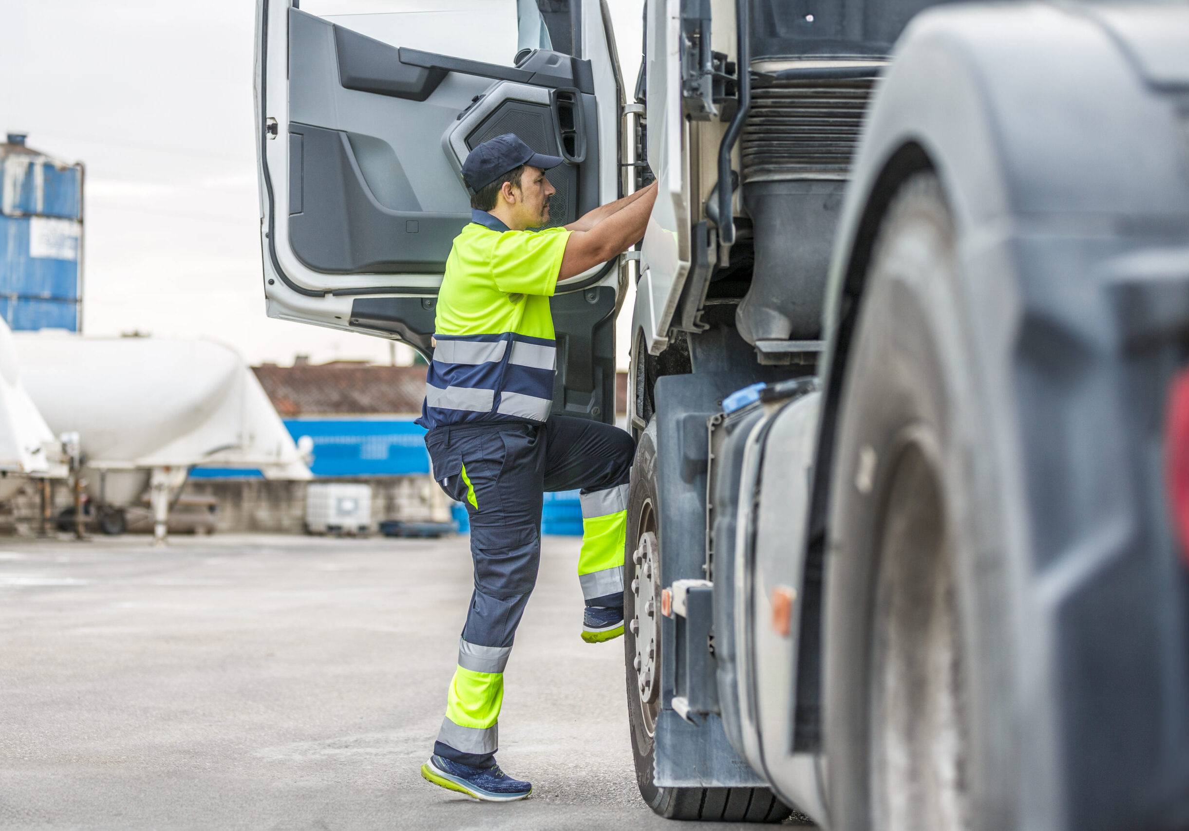Full body side view of focused male driver in uniform and cap climbing steps while entering truck to commence factory delivery operation