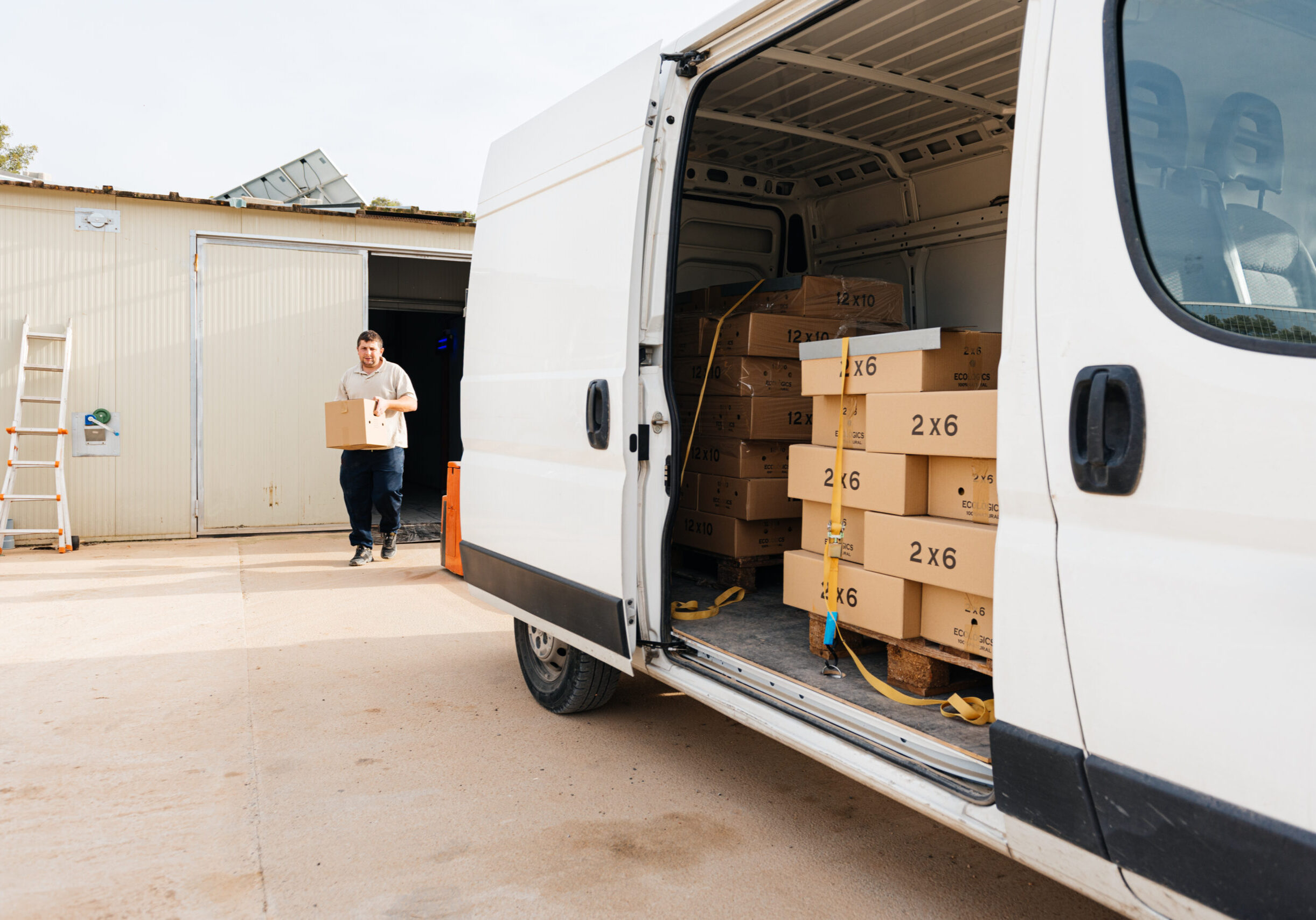 Concentrated young male walking with carton box in hands near warehouse walking towards parked van with open door and loaded cardboard boxes